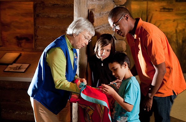 A museum volunteer in a blue vest shows two children and an adult a Seminole blanket in the museum's pioneer cabin.
