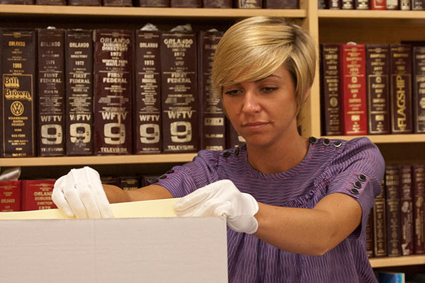 Young lady with short hair wearing gloves looks through box in our 5th floor archives.