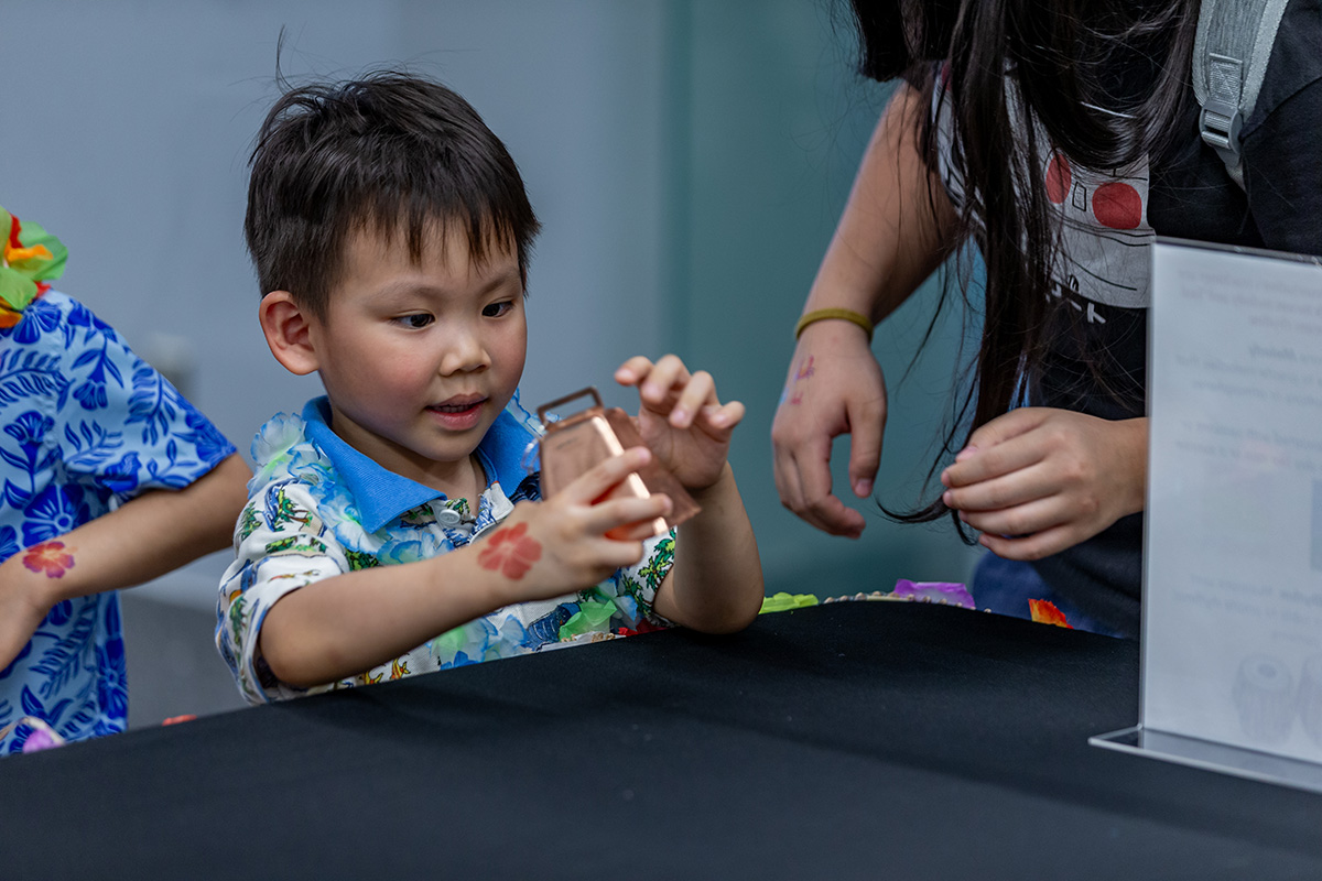 Image of child in blue and white collared shirt inspecting a metal cowbell and a table