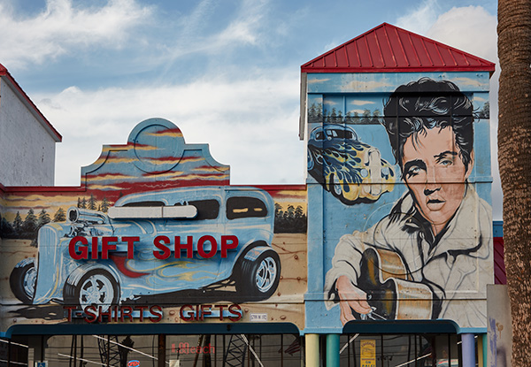 Storefront of the The Wound 'n Around Gift Shop. It features text that says 'Gift Shop' and 'T-shirts and Gifts.' There is a large painting of Elvis holding a guitar and two vintage cars.