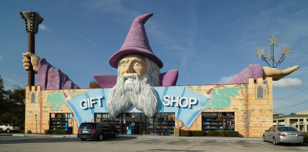 Large building that is designed to look like a castle that says Gift Shop on the front above the doors. Above the building is a large wizard with a purple had and cloaks holding up a staff.